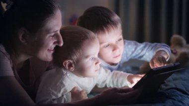 Annesiyle yatakta tablet bilgisayar kullanan pijamalı iki tatlı çocuk. Ailenin birlikte vakit geçirmesi, ebeveynlik, mutlu bir çocukluk ve eğlence.