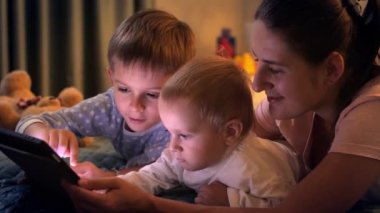 Annesi ve ağabeyi olan bir bebek gece yatağında tablet bilgisayarında oyun oynuyor. Ailenin birlikte vakit geçirmesi, ebeveynlik, mutlu bir çocukluk ve eğlence.