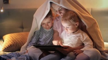 Geceleri battaniyenin altında oturup tablet bilgisayar kullanan mutlu bir aile. Ailenin birlikte vakit geçirmesi, ebeveynlik, mutlu bir çocukluk ve eğlence.
