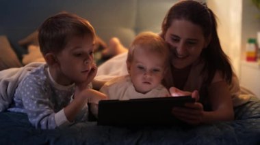 Anne ve çocuklar gece yatağında yatar ve tablet bilgisayarda oyun oynarlar. Ailenin birlikte vakit geçirmesi, ebeveynlik, mutlu bir çocukluk ve eğlence.