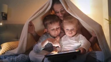 Geceleri battaniyenin altına saklanıp tablet bilgisayar kullanan gülümseyen bir aile. Ailenin birlikte vakit geçirmesi, ebeveynlik, mutlu bir çocukluk ve eğlence.
