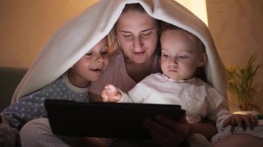 Pijamalı neşeli çocuklar tablet bilgisayarda annelerine oyun oynuyorlar. Ailenin birlikte vakit geçirmesi, ebeveynlik, mutlu bir çocukluk ve eğlence.