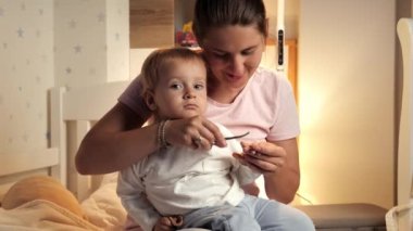 Genç anne yatak odasında küçük oğluna tırnaklarını kesiyor. Ebeveynlik, çocuk hijyeni ve sağlık hizmetleri, uykuya dalmak.