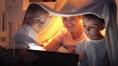 Pijamalı küçük bir çocuk gece tablet bilgisayarında ailesiyle oynuyor. Ailenin birlikte vakit geçirmesi, aletli çocuklar, ebeveynlik, mutlu bir çocukluk ve eğlence.