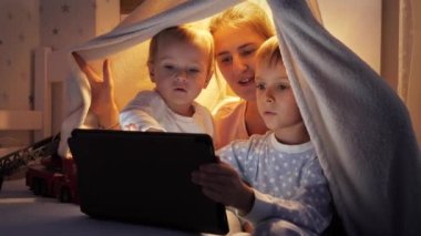 Yatakta çadırda saklanan ve tablet bilgisayarda video izleyen pijamalı gülümseyen aile portresi. Ailenin birlikte vakit geçirmesi, aletli çocuklar, ebeveynlik, mutlu bir çocukluk ve eğlence.