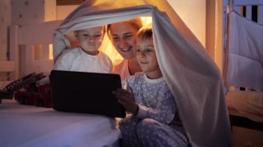 Pijamalı iki gülümseyen çocuğun portresi. Annemle birlikte gece tablet bilgisayardan video izliyorlar. Ailenin birlikte vakit geçirmesi, aletli çocuklar, ebeveynlik, mutlu bir çocukluk ve eğlence.