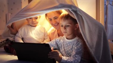 Yatakta battaniye çadırında oturan ve tablet bilgisayar kullanan iki çocukla gülümseyen anne. Ailenin birlikte vakit geçirmesi, aletli çocuklar, ebeveynlik, mutlu bir çocukluk ve eğlence.