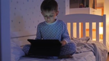 Geceleri pijamalı küçük bir çocuk yatakta oturup tablet bilgisayar kullanıyor. Çocukların eğitimi, gelişimi, alet kullanan çocuklar gizlilik, mahremiyet.