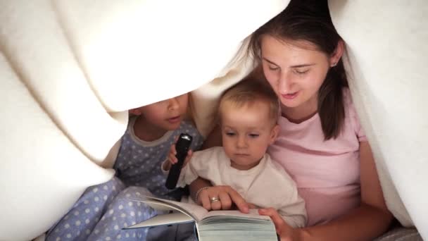 2人の息子の夜に毛布の下に懐中電灯でおとぎ話の本を読んで若い母親 一緒に時間を過ごす家族 子育て 幸せな子供時代とエンターテイメント — ストック動画