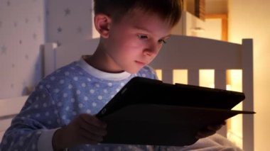 Mutlu gülümseyen çocuğun portresi akşam yatak odasında oturuyor ve tablet bilgisayarda internete bakıyor. Çocukların eğitimi, gelişimi, alet kullanan çocuklar gizlilik, mahremiyet.