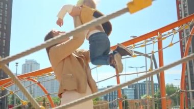 Mutlu anne oğlunu oyun parkında kaldırıyor. Çocuklar dışarıda oynuyor, çocuklar dışarıda, aile yaz tatili ve tatil.