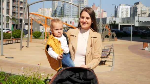 美丽的微笑着的母亲牵着她的小儿子 带着婴儿车在操场上散步 动作缓慢 快乐的养育 有时间在一起的家庭 孩子和父母在户外 — 图库视频影像