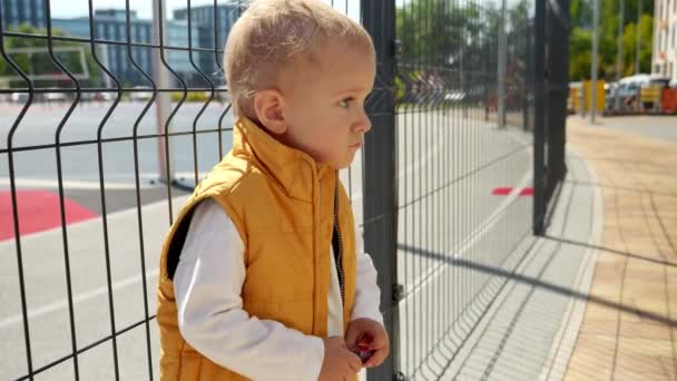 小男孩靠在操场上的金属网栅栏上 烦躁不安的孩子 负面的情绪 孩子的问题 — 图库视频影像