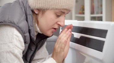 Şapkalı kadın soğuk evde elektrikli ısıtıcıda ellerini ısıtıyor. Enerji krizi, yüksek faturalar, ekonomi ve aylık kamu hizmetleri ödemelerinde tasarruf kavramı