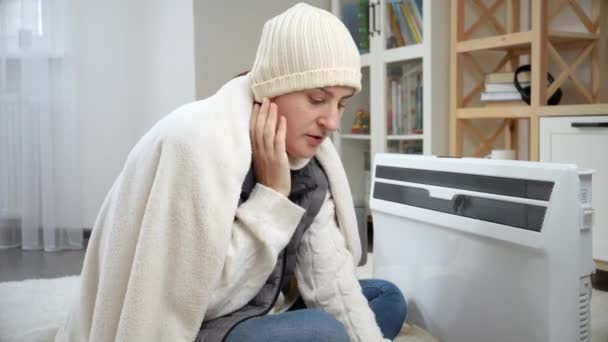 Mujer Joven Sintiéndose Fría Sentada Junto Calentador Eléctrico Poniéndose Sombrero — Vídeos de Stock