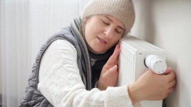Eşarplı ve şapkalı genç bir kadın ısıtıcı kaloriferi kucaklıyor. Enerji krizi, yüksek faturalar, bozuk ısıtma sistemi, ekonomi ve aylık kamu hizmetleri ödemelerinde tasarruf kavramı