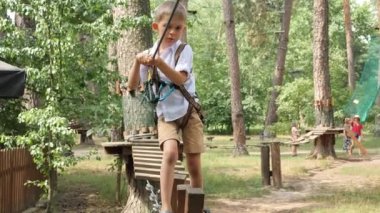 Küçük çocuk sallanan ip köprüde yürüyor ve sıkı bir güvenlik ipi tutuyor. Aktif çocukluk, sağlıklı yaşam tarzı, açık havada oynayan çocuklar, doğadaki çocuklar.