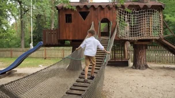 公園の遊び場で木製の吊り橋を歩いている幸せな笑顔の少年 キッズスポーツ 夏休み 屋外での楽しい スカウト — ストック動画