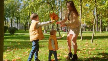 İki mutlu çocuk sonbahar yapraklarını topluyor ve parkta annelerine veriyorlar. Aile açık havada, mutlu ebeveynlik ve çocukluk, sonbahar manzarası
