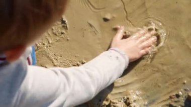 Sahilde oturan ve elleriyle kum kazan küçük çocuğun yakın çekimi. Çocuk dışarıda oynuyor, çocuklar sahilde, gölde dinleniyor..