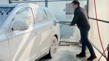 Genç adam arabasını köpükle ve araba yıkamada fırçayla yıkıyor. Otomobil bakımı, ulaşım temizliği, kirli araba.