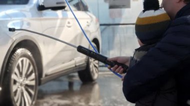 Bebek bebekle birlikte araba yıkamada su püskürterek araba yıkayan bir adam. Otomobil bakımı, ulaşım temizliği, kirli araba.