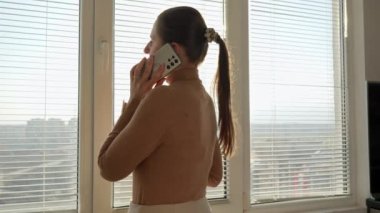 Pencerenin önünde panjurları olan ve telefonla konuşan güzel bir kadın. Sabahları insanlar, güzel şehir manzarası, ofiste mola vermek.