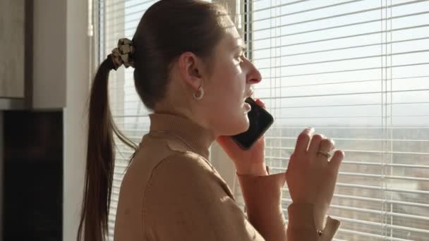 Młoda Zmartwiona Kobieta Rozmawiająca Przez Telefon Zaglądająca Przez Okno Świadek — Wideo stockowe