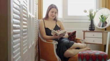 Gülümseyen zarif bir kadın yatak odasında koltukta otururken kitap okuyor. Kadın eğlencesi, eğitim, evde rahatlama