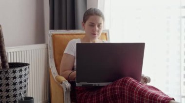 Pijamalı genç bir kadın büyük pencerede dizüstü bilgisayarla çalışıyor. Evde çalışan serbest çalışan, uzaktan çalışan, evde iş yapan bir kadın.