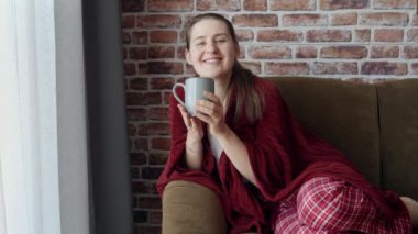 Pijamalı, ekoseli ve kanepede çay içen mutlu bir kadın. Kadın evde dinleniyor, yaşam tarzı ve rahatlama.