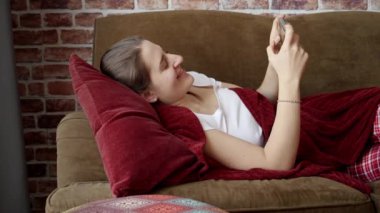 Oturma odasındaki kanepede dinlenen ve akıllı telefon kullanan genç bir kadın. Evde dinlenen insanlar, iletişim, uzaktan çalışma