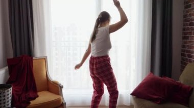 Oturma odasındaki büyük pencerede pijamalı komik bir kadın dans ediyor. İnsanlar evde rahatlıyor, eğleniyor, olumlu duygular ve eğlence yaşıyor.