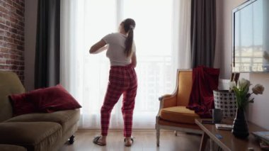 Mutlu bir kadının pijama giyip oturma odasındaki büyük pencerenin önünde dans ettiği komik bir fotoğraf. İnsanlar evde rahatlıyor, eğleniyor, olumlu duygular ve eğlence yaşıyor.