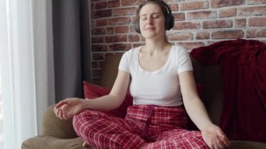 Kulaklıkla mantraları dinleyen ve evde meditasyon yapan genç bir kadın. Rahatlama, sağlıklı yaşam tarzı, evde dinlenme ve yoga yapma kavramı.