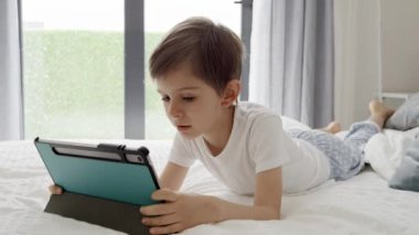 Genç çocuk dijital dünyayı keşfetmek için bir tablet bilgisayar kullanarak yatağında ekranda zaman geçirmekten hoşlanıyor. Alet, eğitim ve geliştirme kullanan çocuk