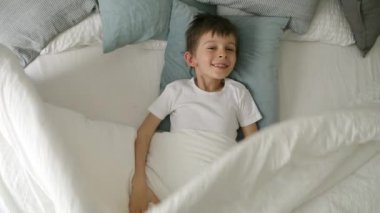 Neşeli bir çocuk yatakta yatar ve kameraya yöneltilmiş bir gülümsemeyle battaniyesini açar, neşe ve neşe dolu bir ruh gösterir.