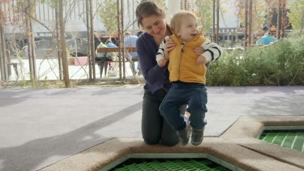 幸せな陽気な赤ちゃん男の子とともに母ジャンプオントランポリンで遊び場で公園 — ストック動画