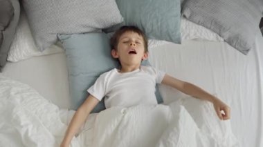 Mutlu gülümseyen çocuk battaniyesini açar ve yatakta bir sürü yumuşak yastıkla uzanır..