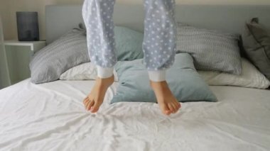 Yatak odasındaki yumuşak yatakta yükseğe zıplayan bir çocuğun yavaş çekimi. Mutlu çocukluk, eğlence ve neşe
