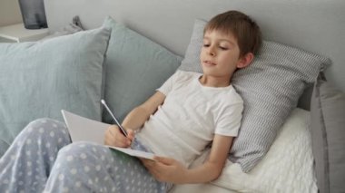 Genç bir çocuk yatakta uzanıyor, ödevini bir defterde yapıyor. Eğitimin önemi ve çocuk gelişimi, evde uzaktan eğitim.