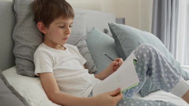 Küçük çocuk yatakta ödev yapıyor, bir deftere yazıyor. Eğitim, çocuk gelişimi ve evden uzak eğitim kavramı