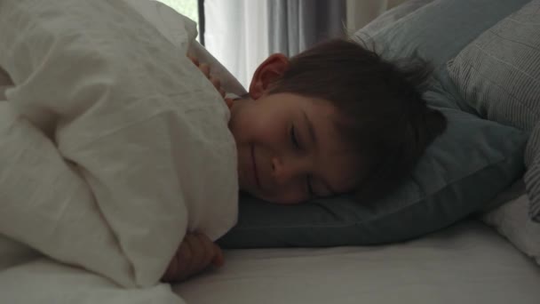 窓が開いて太陽が窓を通して輝くようにベッドで目を覚ます笑顔の男の子の肖像画 — ストック動画