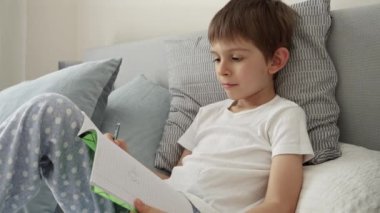 Genç çocuk yatakta yatarken defterine yazıyor. Eğitimin özü, çocuk gelişimi ve ev ortamında uzaktan öğrenme