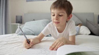 Yatakta ödevlerini yaparken pijamalarıyla deftere yazı yazan genç bir çocuk. Uzak bir öğrenim ortamında eğitim ve çocuk gelişimi