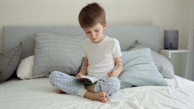 Küçük çocuk yatakta oturuyor ve kurşun kalemle defterine yazıyor. Evde eğitim, çocuk gelişimi ve uzaktan eğitim kavramı