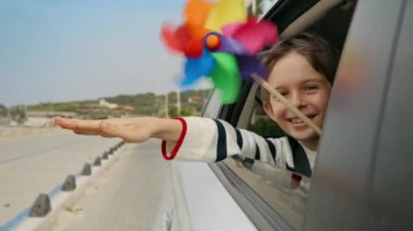 Araba camından dışarı bakarken elinde renkli bir değirmen ya da değirmen tutan mutlu çocuk. Çocuk seyahati, seyahat, turizm, tatil, hafta sonu ve tatil kavramı