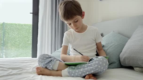 パジャマ姿の可愛い男の子がベッドに座ってノートに宿題をしている 家庭での教育 子供の発達 遠隔教育の概念 — ストック動画