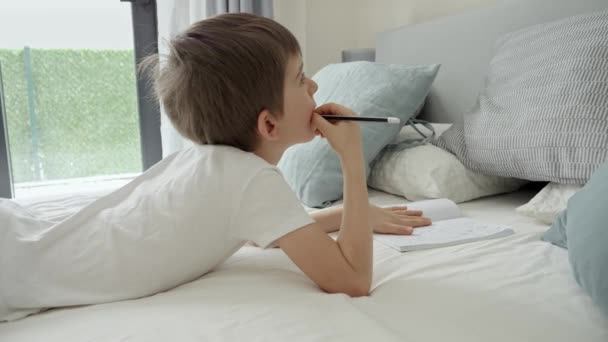 朝起きてベッドに横になって宿題をしている可愛い男の子 家庭での教育 子供の発達 遠隔教育の概念 — ストック動画