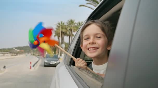 カラフルな回転するピンホイールを持っている陽気な少年は 彼が開いている車の窓の外を眺めると微笑む 子供の旅行 休暇の概念 — ストック動画